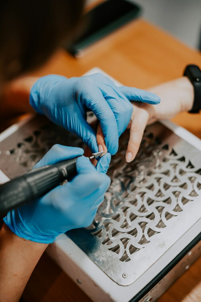 Person wearing blue gloves filing fingernails with an electric nail drill, demonstrating care and precision in nail grooming.