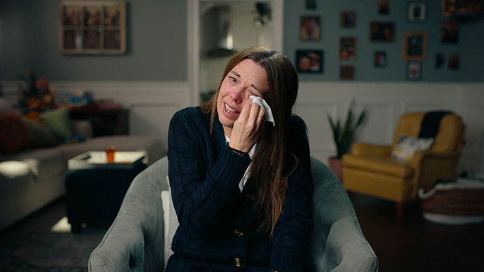 Woman sitting in a living room, wiping tears from her face, reflecting the emotional impact of secret torment by a mom.