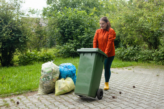 Woman moving a green garbage bin on a driveway near several trash bags in a residential outdoor setting. Woman moving a green garbage bin on a driveway near several trash bags in a residential outdoor setting.