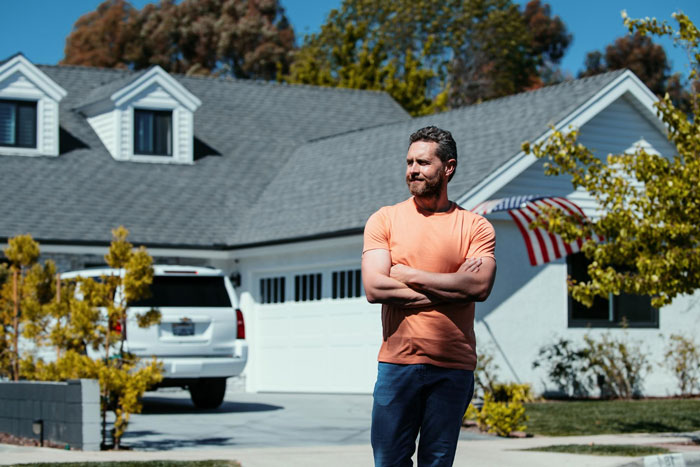 Man standing with arms crossed in front of house driveway under sunny sky, relating to garbage bins and neighbor dispute. Man standing with arms crossed in front of house driveway under sunny sky, relating to garbage bins and neighbor dispute.