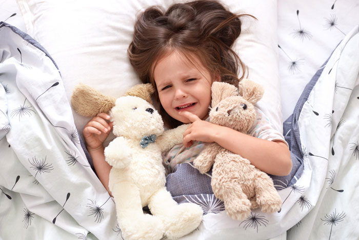 Young girl upset in bed holding stuffed animals, illustrating kid being kicked out of roommate&rsquo;s bed conflict scenario.