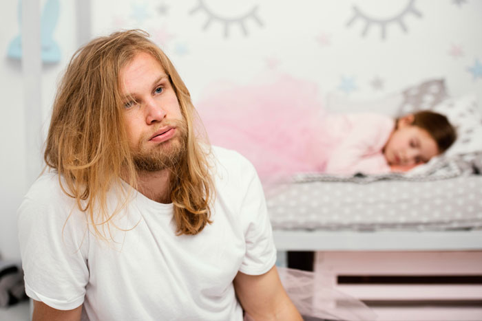 Young man sitting thoughtfully in a room while a child sleeps in a bed, reflecting tension over roommate sleeping arrangements.