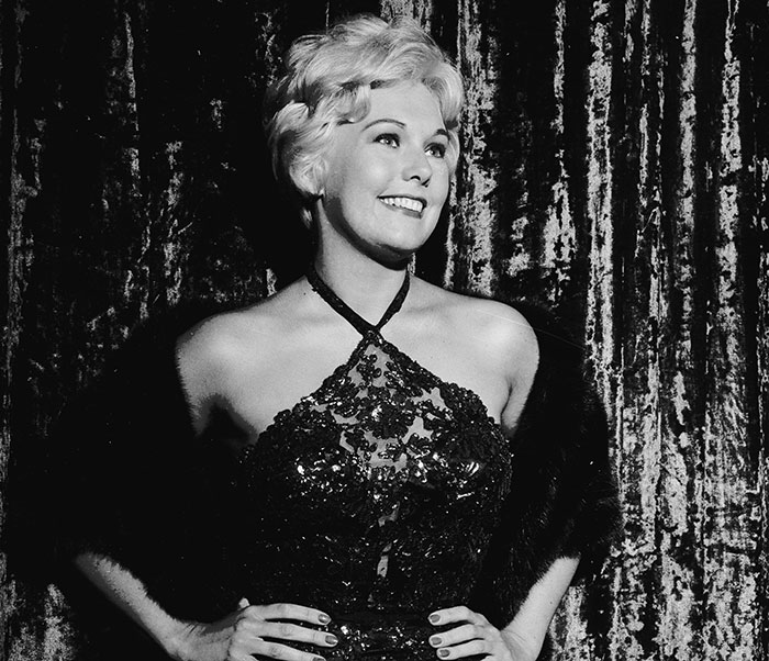 Kim Novak smiling in a glamorous lace dress with a fur stole, receiving a lifetime achievement award at Venice Film Festival.