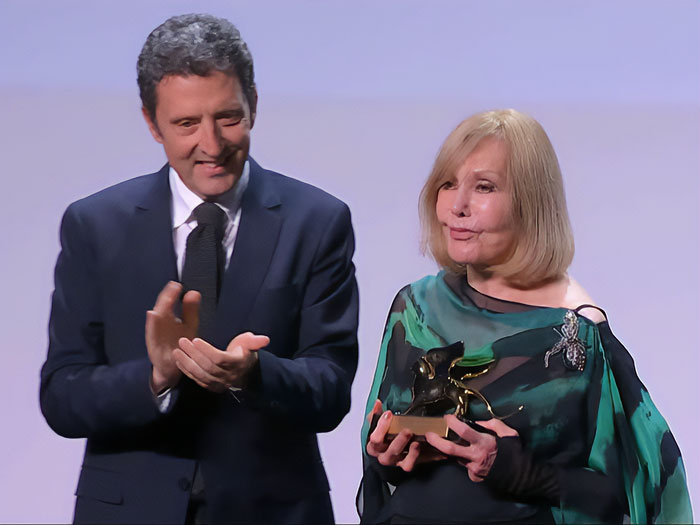 Kim Novak holding a lifetime achievement award at Venice Film Festival while a man in suit applauds beside her.
