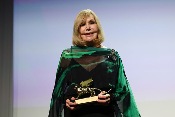 Kim Novak in green and black outfit holding a golden award while posing at Venice Film Festival event.