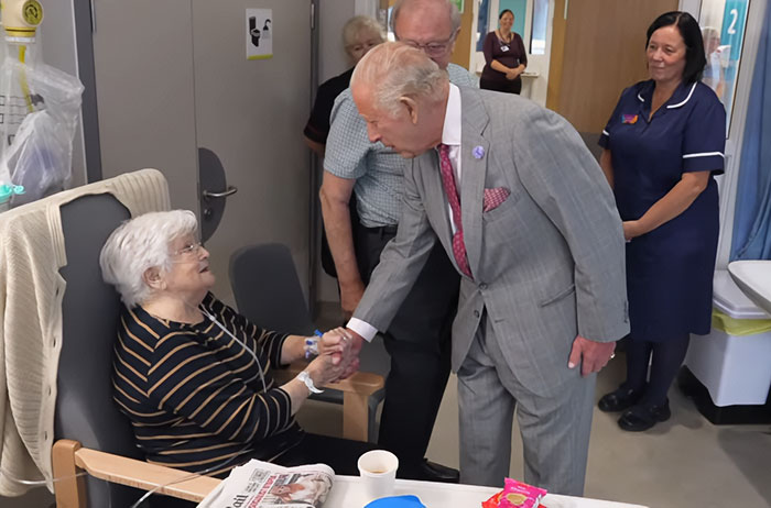 King Charles in a grey suit shaking hands with an elderly woman in a hospital, sharing an update on his incurable cancer.