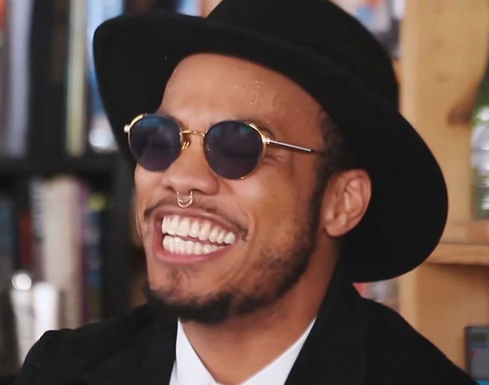 Man wearing round sunglasses and a black hat smiling brightly, featured in stories from people who knew celebrities before fame.