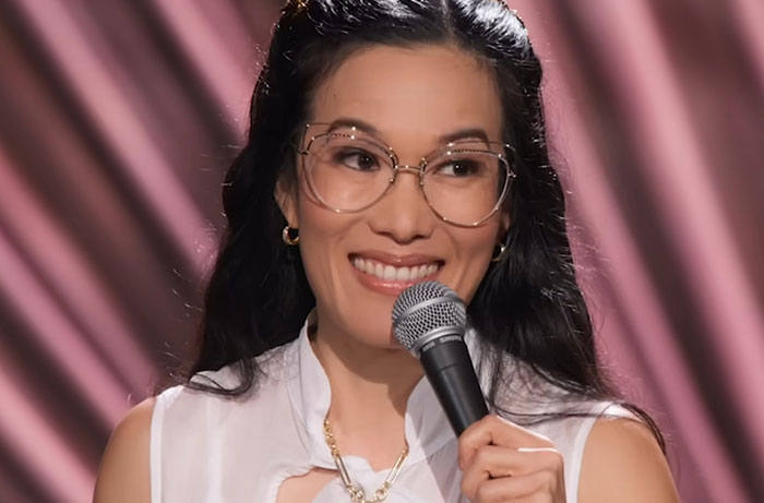Woman with glasses holding a microphone, smiling while sharing stories about celebrities before fame in a stand-up setting.