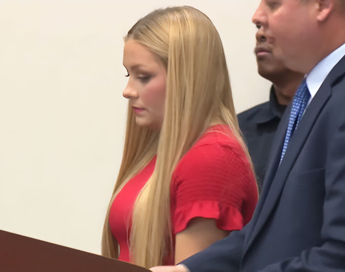 Young woman in a red dress, standing next to men in suits, related to cheerleader baby belly case in court setting. Young woman in a red dress, standing next to men in suits, related to cheerleader baby belly case in court setting.