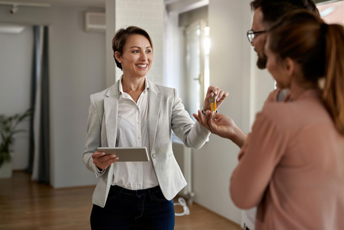 Smiling woman handing over keys to a couple in a bright room, illustrating landlord malicious compliance drama concept.