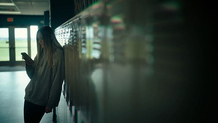 Teen girl leaning against school lockers, looking at phone, illustrating themes of secret torment and emotional distress.