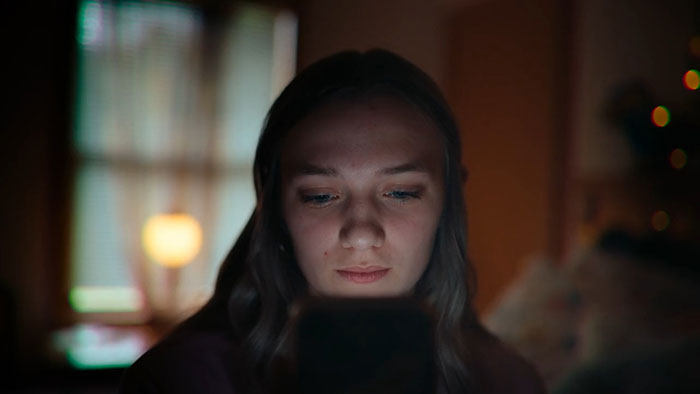 Teen girl looking at her phone with a somber expression in a dimly lit room, highlighting emotional impact of bullying.