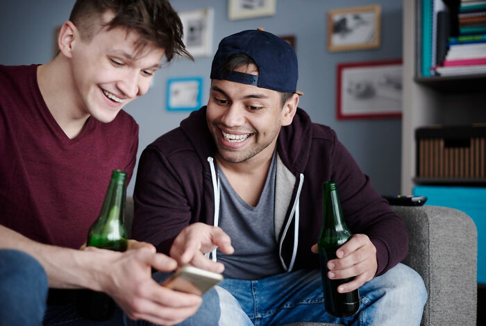 Two young men laughing and holding beer bottles while looking at a smartphone, illustrating dumb ways folks incriminated themselves.