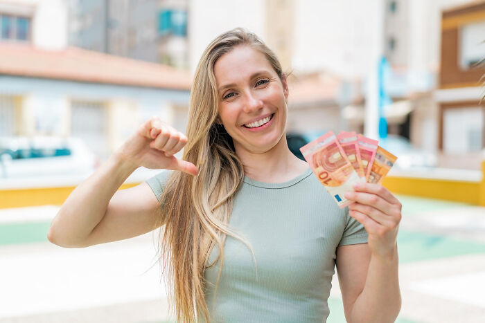 Woman smiling outdoors, holding cash in hand and pointing at herself, illustrating dumb ways people incriminated themselves.