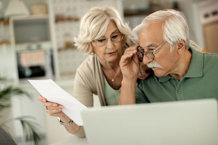 Elderly couple uncovering darkest family secrets, looking concerned while reviewing documents and laptop together at home.