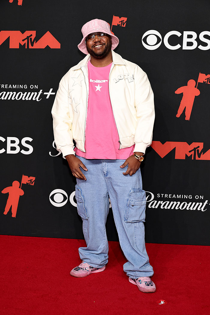 Man wearing pink bucket hat, pink shirt, cream jacket, and baggy jeans posing on red carpet at worst dressed stars 2025 Video Music Awards.