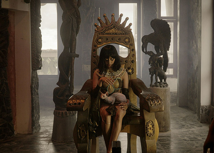 Woman dressed in ancient attire sitting on a throne holding a hairless cat in a dim, eerie room with sculptures.