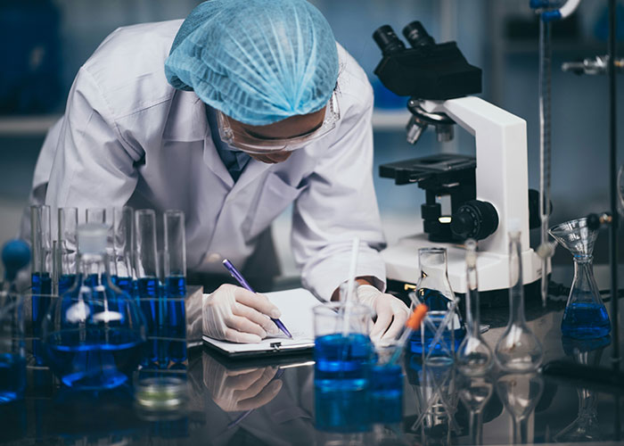 Scientist in a lab coat and hairnet recording data surrounded by test tubes and a microscope in a scientific setting