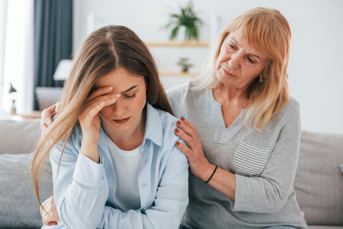 Creepy MIL comforting upset pregnant DIL who looks stressed while sitting on a couch in a cozy living room.