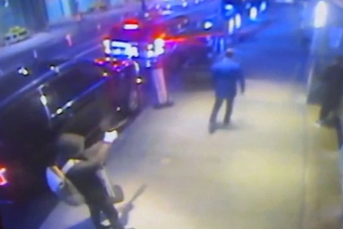 Surveillance footage showing a person bending near a vehicle as police cars with flashing lights arrive on the street.