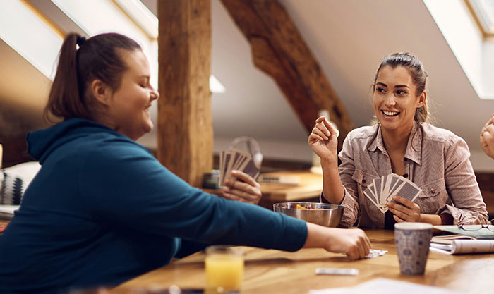 Two women enjoying a card game at a wooden table, smiling and engaging in a relaxed indoor setting for lying pick-me girl tattoo inspiration.