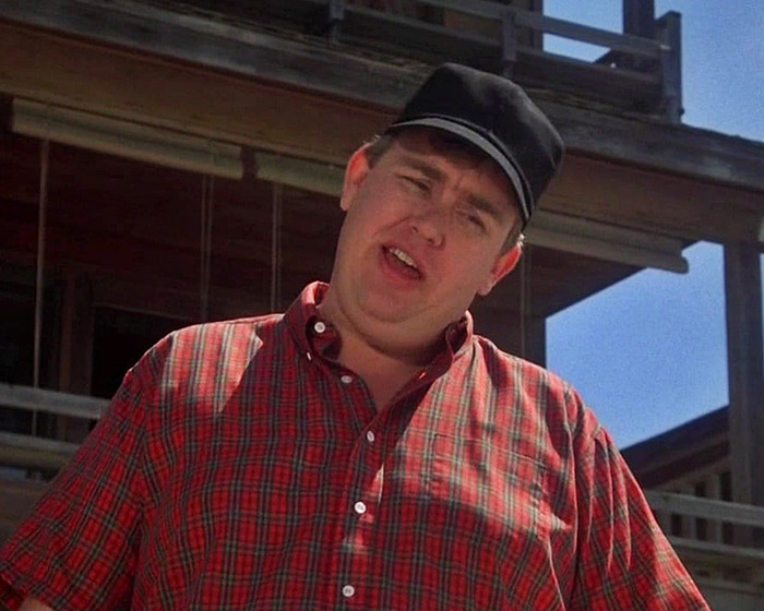 John Candy in a red plaid shirt and black cap, outdoors in front of a wooden structure on a sunny day. John Candy in a red plaid shirt and black cap, outdoors in front of a wooden structure on a sunny day.
