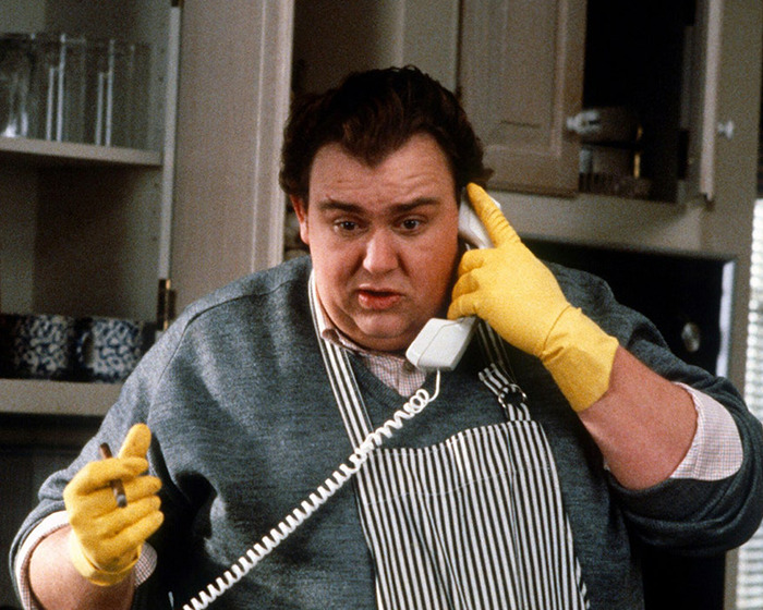 John Candy wearing yellow gloves and apron, holding a phone receiver in a kitchen setting. John Candy wearing yellow gloves and apron, holding a phone receiver in a kitchen setting.