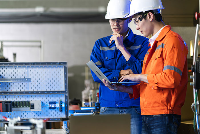 Two engineers wearing safety helmets and workwear, discussing a project on a laptop in an industrial setting.