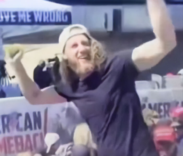 Bearded man in crowd cheering and looking back during Charlie Kirk's attack, captured with raised arms and a smile.
