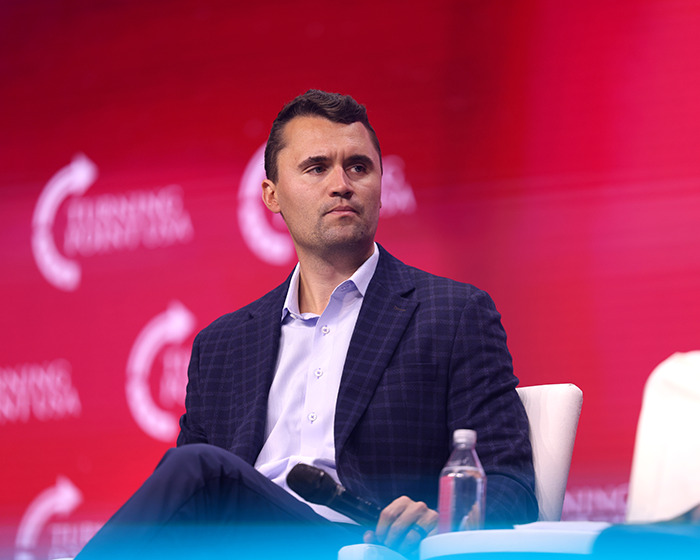 Man sitting on stage wearing a blue checkered suit, representing Charlie Kirk in a public speaking event.