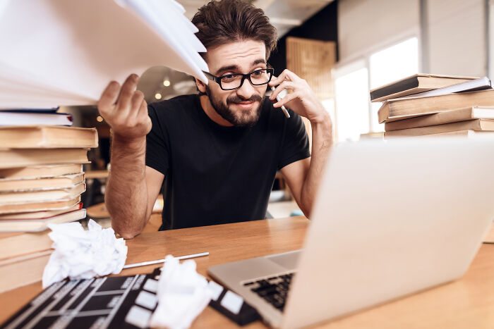 Man holding papers and talking on phone while working on laptop surrounded by stacks of books, illustrating employee insider tricks.