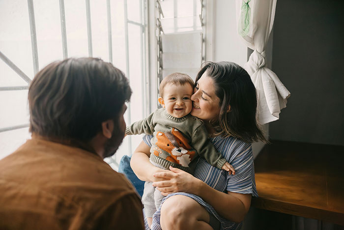 Mother holding baby smiling at man by window, illustrating robotically calculated choice based on brains over heart.