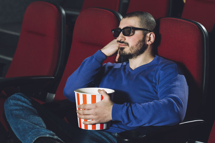 Man in sunglasses sitting alone in theater holding popcorn, symbolizing tests women use to spot red flags on dates.