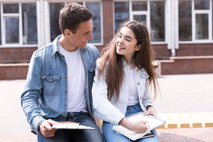 Young couple sitting outdoors with notebooks, discussing an art class memory disagreement between them.