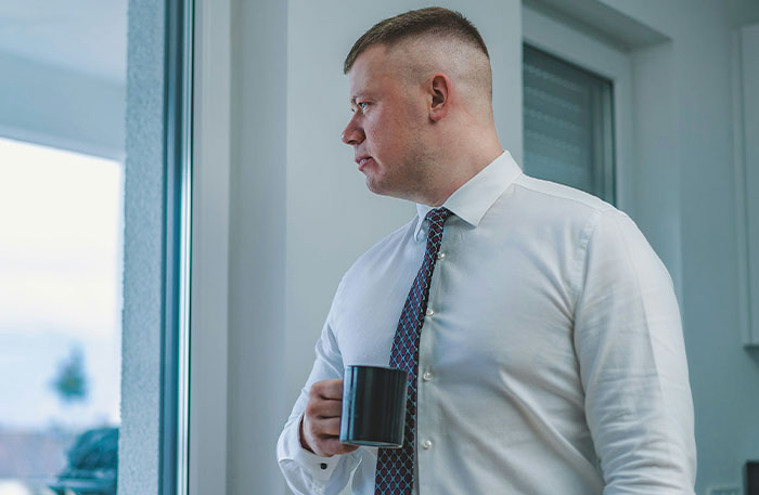 Employee in white shirt and patterned tie looks out window holding black mug, expressing frustration related to promised raises.