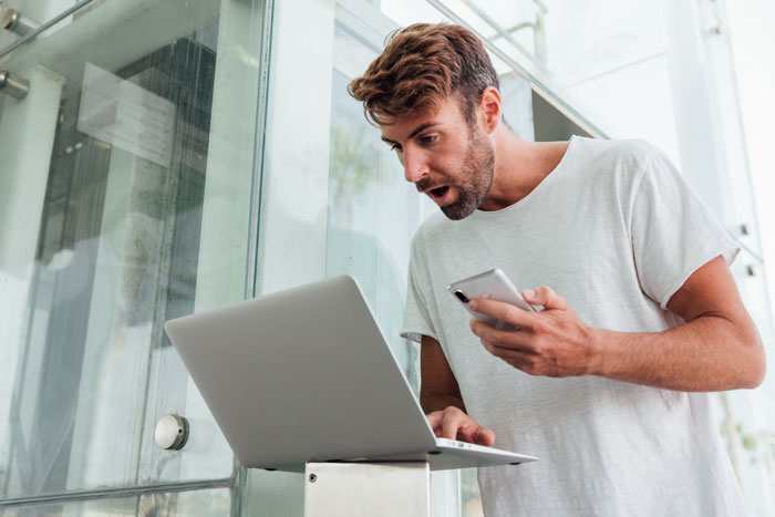 Man reacting with surprise at laptop while holding phone, illustrating employees maliciously complying with strict dress code. Man reacting with surprise at laptop while holding phone, illustrating employees maliciously complying with strict dress code.