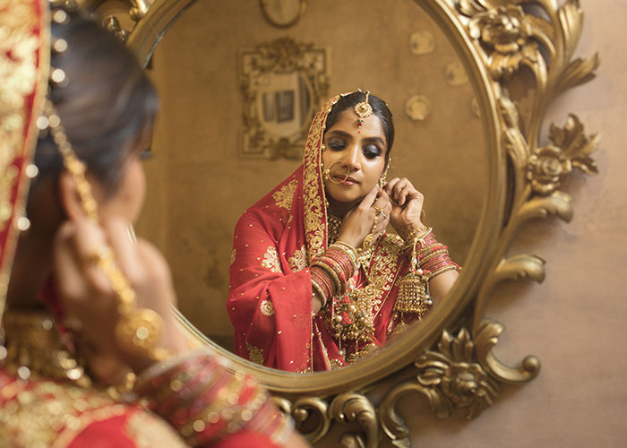 Woman in traditional bridal attire adjusting jewelry in front of an ornate mirror at a wedding event.