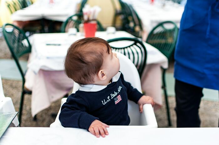 Baby sitting in a high chair at a restaurant, looking up towards a server, capturing a candid dining moment.
