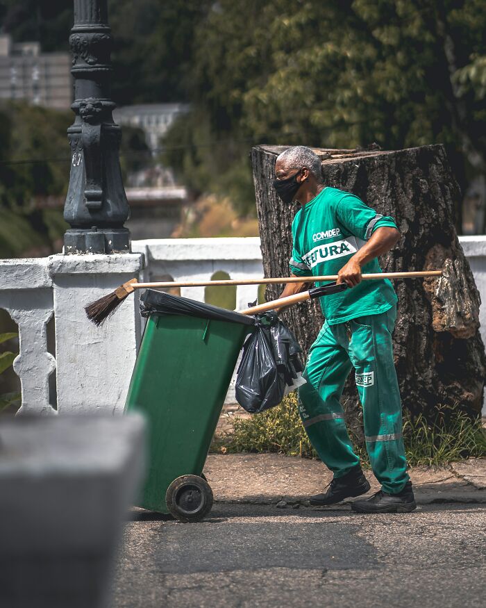 Man in green uniform and black mask pushing a trash bin and broom while cleaning outdoors in the Bahamas street.