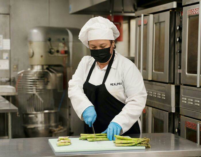 Woman chef wearing a mask and gloves cutting asparagus in a commercial kitchen, illustrating subtle misogyny in daily lives.