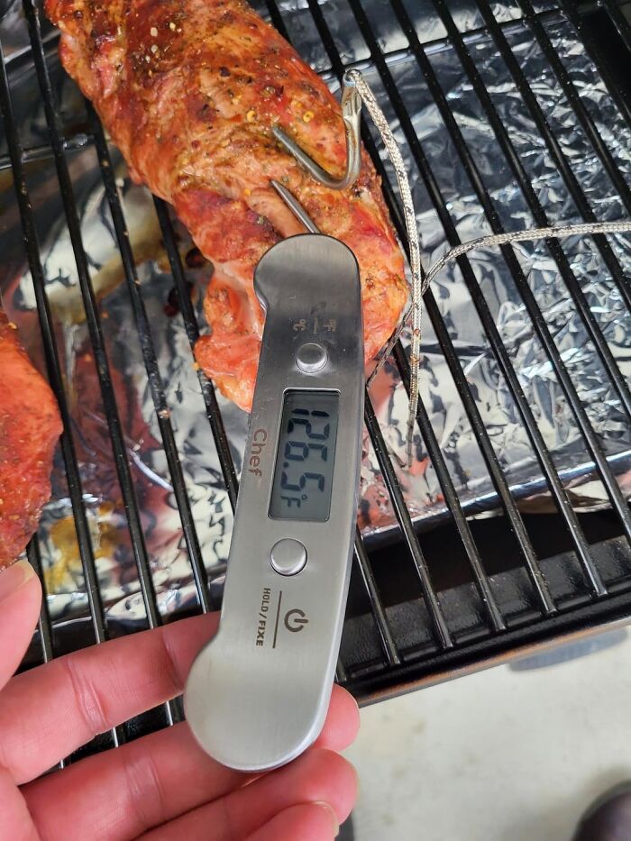 Hand holding digital meat thermometer reading 126.5°F inserted into grilled meat on a barbecue grill, kitchen tricks.