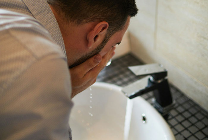 Man washing face over bathroom sink, illustrating downplayed medical conditions and procedures affecting men.