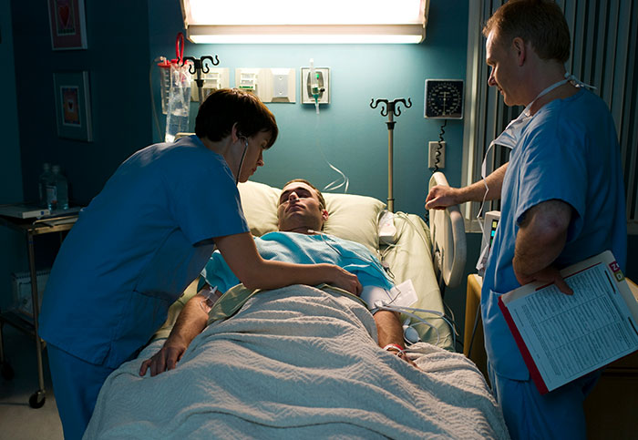 Male patient in hospital bed monitored by two nurses, highlighting downplayed medical conditions and procedures.