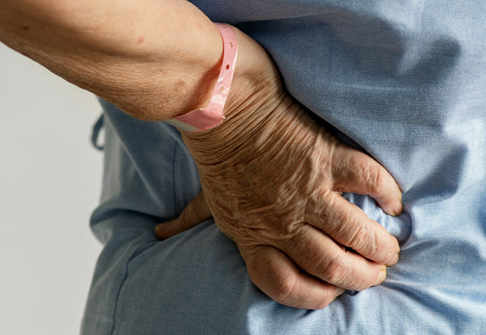 Elderly person wearing hospital wristband holding lower back, illustrating downplayed medical conditions and procedures.