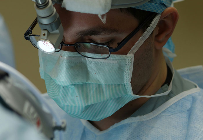 Surgeon wearing mask and glasses focused on medical procedure highlighting downplayed medical conditions and procedures.