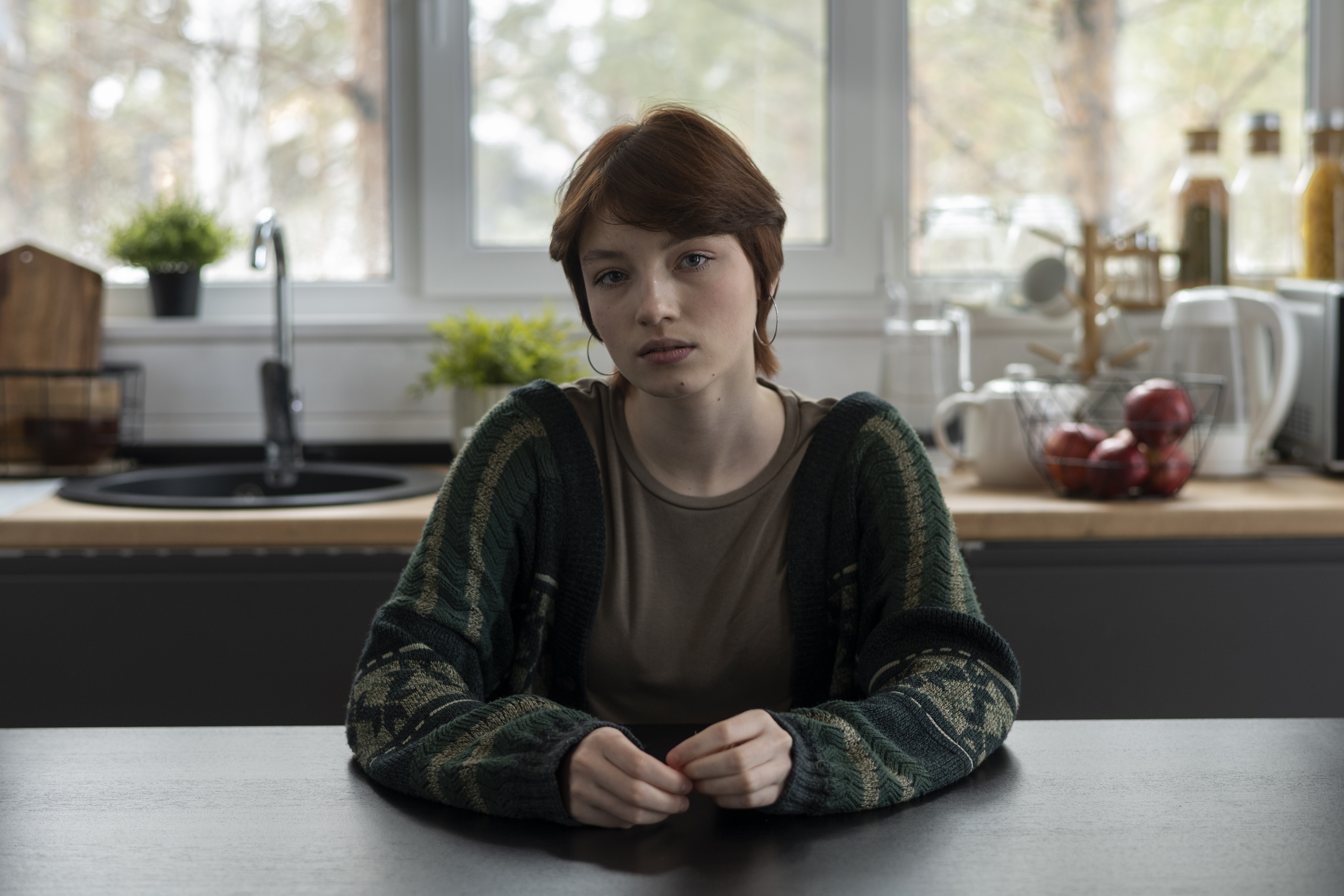 Young woman sitting at kitchen table, looking upset as family demands she gives her baby to her sister.