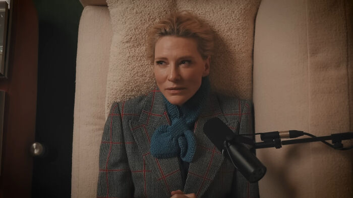 Woman in a plaid blazer and blue scarf speaking into a microphone during a heartwarming celebrity encounter podcast.