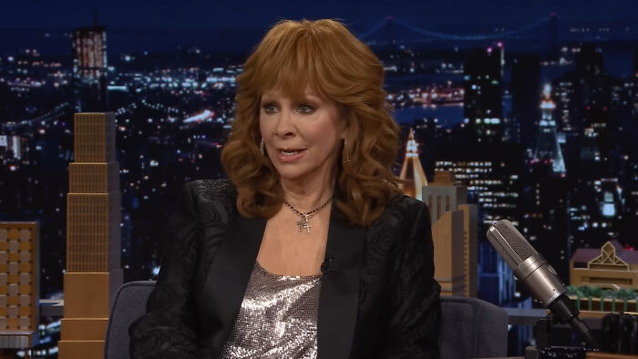Reba McEntire in a TV interview wearing a sparkly top and black jacket, sharing heartwarming celebrity encounter stories.