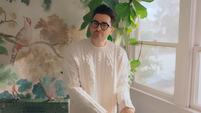 Man wearing glasses and a white sweater posing indoors with plants and wallpaper reflecting heartwarming celebrity encounters.