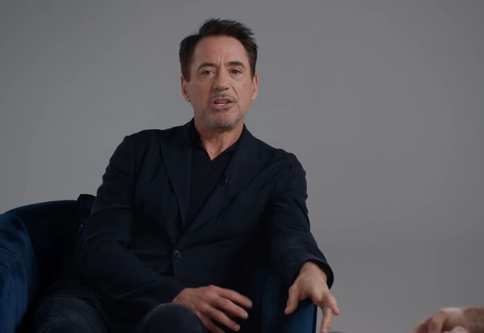 Man in dark blazer sharing a heartwarming celebrity encounter during a casual interview setting with a gray background.